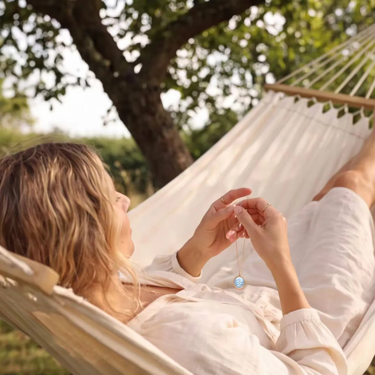 Blonde woman relaxing outdoors on a hammock while admiring a kawaii zodiac necklace with iridescent mother-of-pearl pendant by OceanCarvingLab.