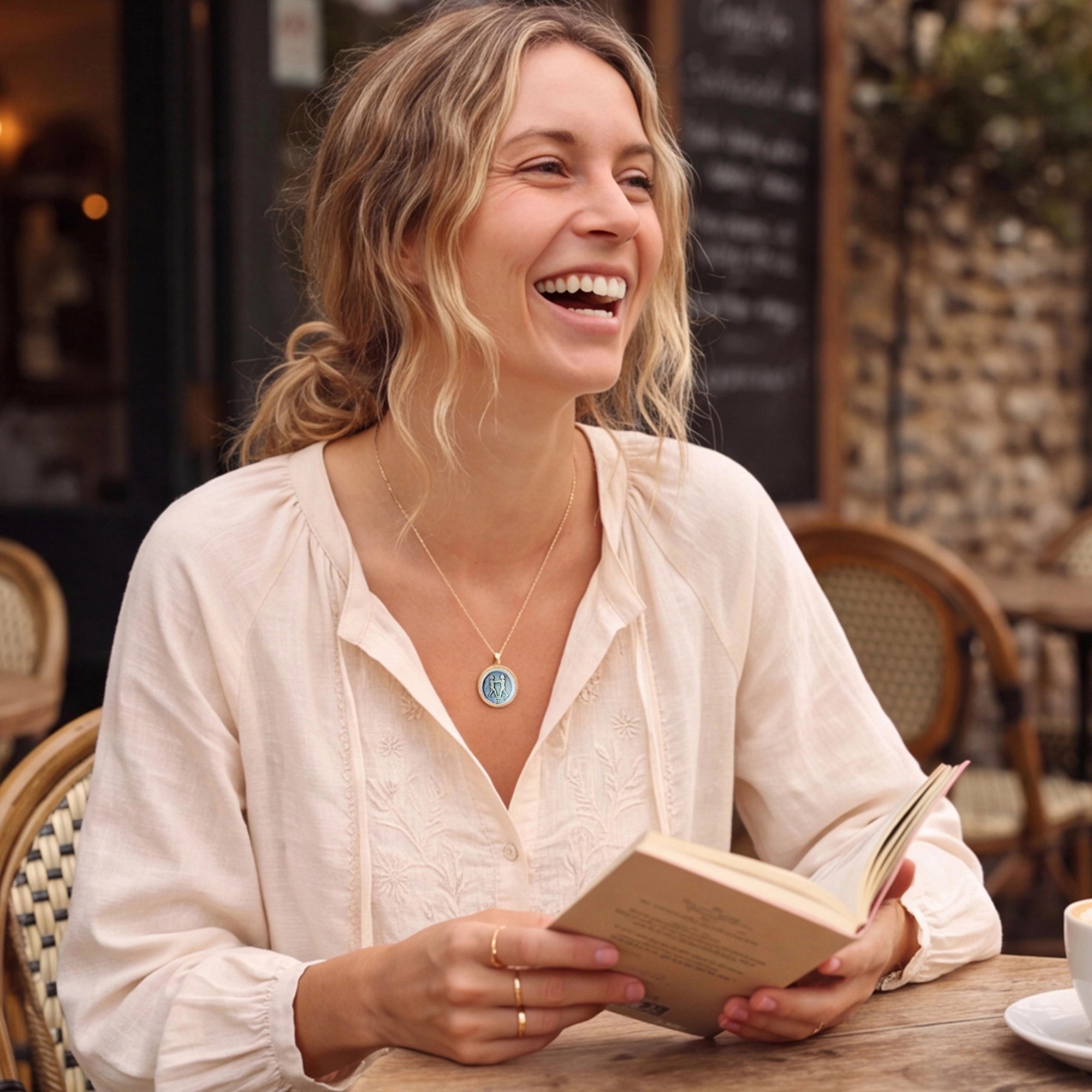 Elegant blonde woman reading outdoors wearing a Van Gogh style carved zodiac mother-of-pearl pendant necklace in gold plated sterling silver by OceanCarvingLab.